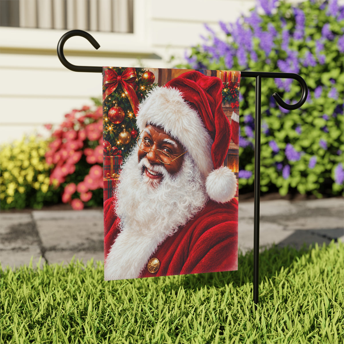 A rectangular Christmas garden flag featuring a festive portrait of a black Santa Claus, displayed on a black metal garden stake in a grassy yard with colorful flowers in the background.