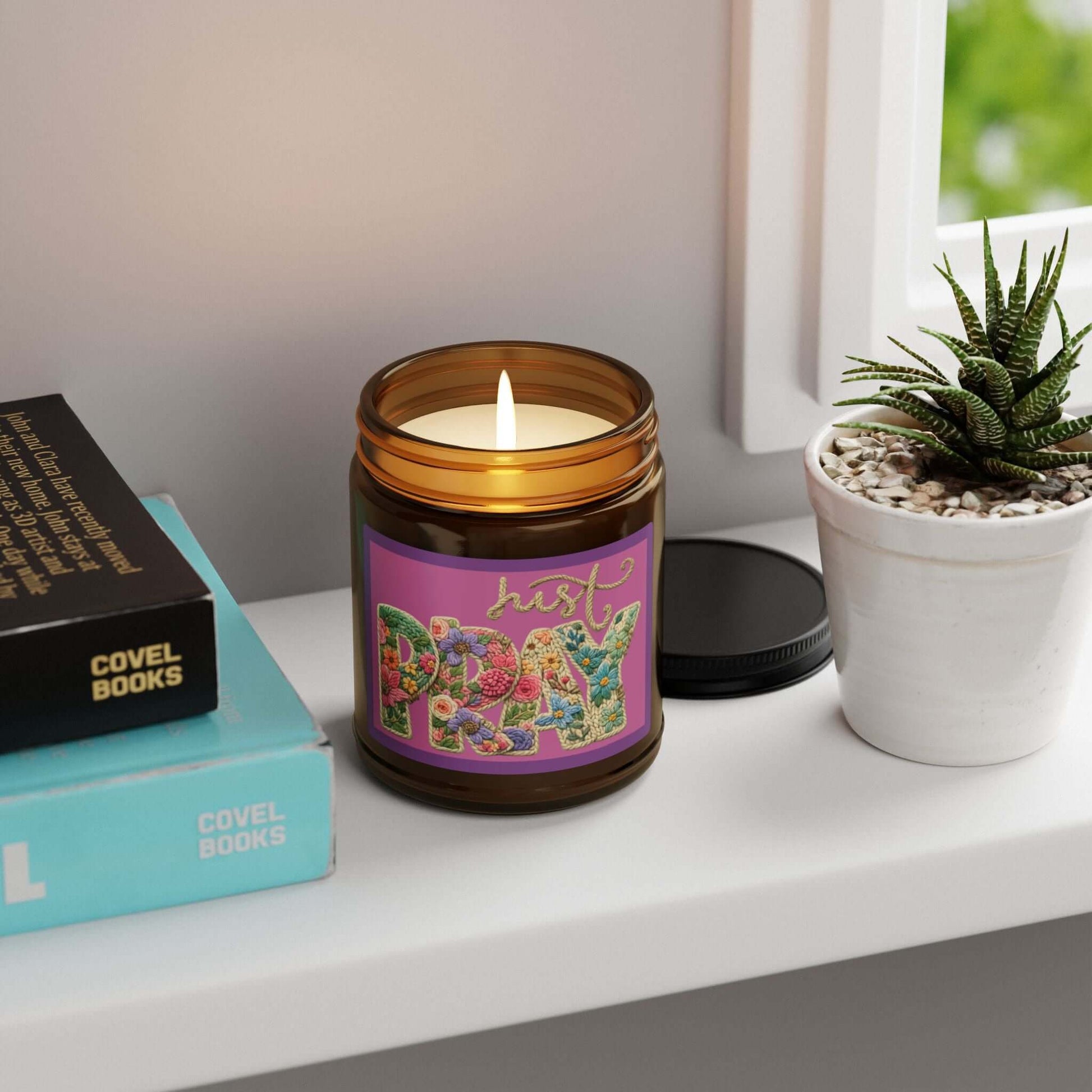 Just Pray Vegan Soy Candle in an amber jar with a warm glow, placed on a shelf next to books and a small plant.