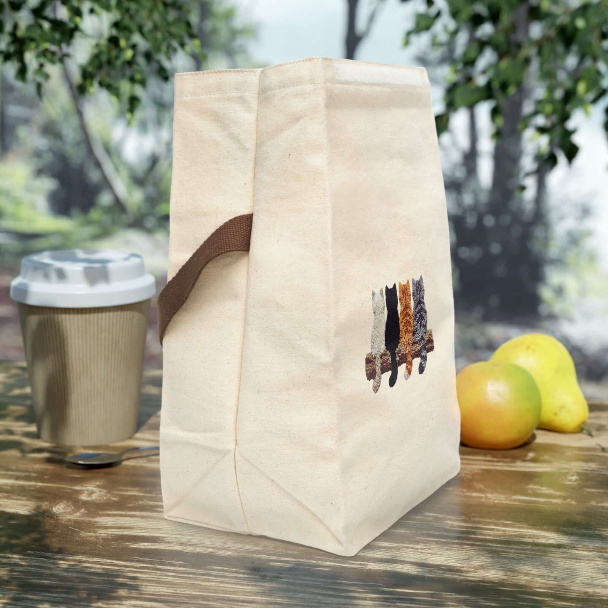 Simply Cute Kittens Canvas Lunch Bag with Strap on a picnic table with a coffee cup and fruits.